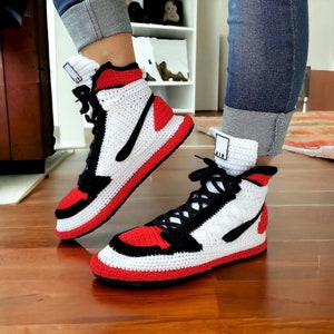 Puede incluir: Zapatillas altas de ganchillo hechas a mano en una combinación de colores clásica. Los zapatos presentan una base blanca con detalles en rojo y negro, incluyendo un swoosh y cordones negros. Las suelas son rojas y los puños del tobillo tienen una pequeña etiqueta blanca con la palabra "AIR".