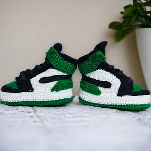 Op de afbeelding: Een paar groene, witte en zwarte gehaakte babyschoenen. De schoenen zijn in de stijl van een populaire basketbalschoen.
