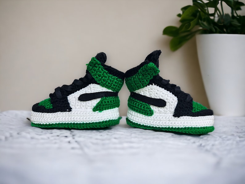Op de afbeelding: Een paar groene, witte en zwarte gehaakte babyschoenen. De schoenen zijn in de stijl van een populaire basketbalschoen.