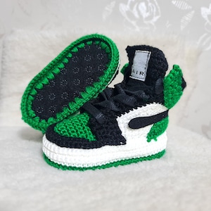 Op de afbeelding: Groene en zwarte gehaakte babyschoenen met witte accenten en het woord "AIR" aan de zijkant.