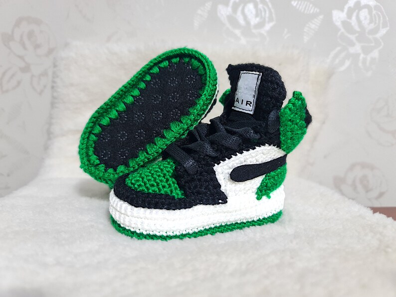 Op de afbeelding: Groene en zwarte gehaakte babyschoenen met witte accenten en het woord "AIR" aan de zijkant.
