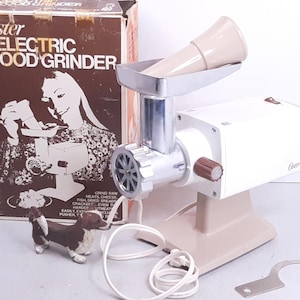 May include: A white Oster electric food grinder with a beige base and a chrome grinder attachment. The grinder has a brown knob and a beige plastic food pusher. The box for the grinder is brown with white text that reads "Oster Electric Food Grinder".