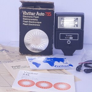 May include: A black Vivitar Auto 215 electronic flash with its original box and paperwork. The box is black with white text and a silver circular design. The flash unit has a rectangular head and a small control panel.