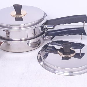 May include: A set of three stainless steel pots with black handles and gold accents. The pots are stacked on top of each other, with the largest pot on the bottom and the smallest pot on top. The pots have lids.