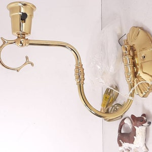 May include: A gold-toned wall sconce with a decorative, curved arm and a floral design. The sconce is attached to a white wall and has a clear plastic bag and a small dog figurine in the foreground.