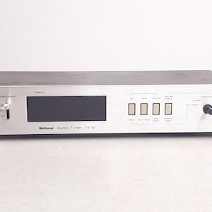 May include: Silver National Audio Timer TE 97 with a black display screen and multiple buttons. The device has a dial for outlet control and several function buttons labeled "Test", "Slow", "Monitor", and "Check".