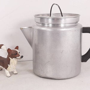 May include: A silver aluminum coffee pot with a black plastic handle and a wire lid. The pot has a textured surface and is sitting on a white surface.