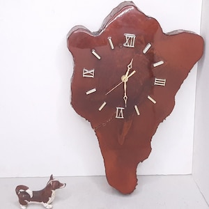 May include: A unique wall clock crafted from a piece of wood, featuring a natural, irregular shape. The clock face is a deep brown with gold-colored hands and Roman numeral hour markers. A small dog figurine is in the foreground.