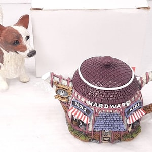 May include: A ceramic teapot shaped like a hardware store with a red roof, white trim, and the words "HARDWARE" in black lettering. The teapot has a red and white striped awning with the words "TOOLS" and "NAILS" on either side.