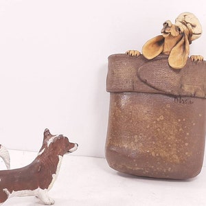 May include: A brown ceramic figurine of a dog peeking out of a brown ceramic boot. The dog has a white hat on its head. A smaller brown and white ceramic dog figurine is standing in front of the boot.