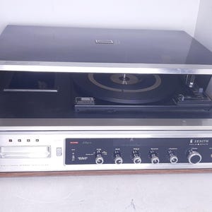 May include: Vintage Zenith solid-state stereo console record player. The unit has a black top and a silver and wood-tone front panel with various controls and a cassette player. The turntable is visible inside.