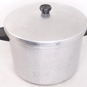 May include: A large, round, aluminum pot with a lid and two black handles. The lid has a black knob. The pot is designed for cooking and has a simple, utilitarian design. The pot is likely used for cooking large quantities of food.