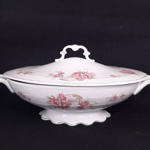 May include: White porcelain serving dish with a lid, adorned with pink rose floral patterns and gold trim. Features ornate handles and a decorative base. A vintage piece for serving food.