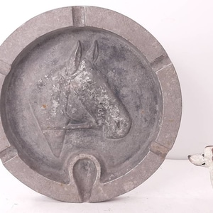 May include: A gray metal ashtray with a horse head design and a horseshoe cutout. The ashtray is round and has a raised rim.
