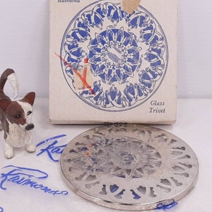 May include: A silver-colored glass trivet with a decorative design. The trivet is round and has a cut-out design. The trivet is packaged in a white box with the words "Glass Trivet" printed on it.