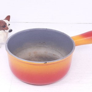 May include: A small, round cooking pot with a single handle. The pot is painted in a gradient of colors, transitioning from red at the base to yellow at the handle. The interior is a dark gray color. A decorative dog figurine is in the background.