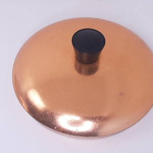 May include: A copper-colored lid with a black knob. The lid is round and has a shiny, reflective surface. The knob is cylindrical and sits in the center of the lid. The lid appears to be made of metal.