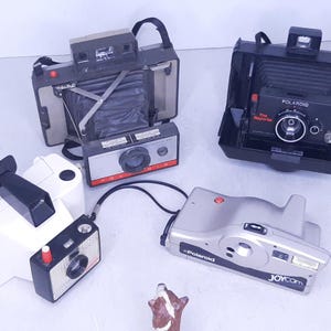 May include: Four vintage Polaroid cameras, including a black and silver Polaroid Land Camera, a black Polaroid The Reporter camera, a white Polaroid camera with a Joycam lens, and a silver Polaroid camera with a red button.