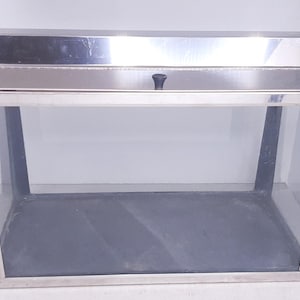 May include: A rectangular display case with a stainless steel frame and a glass enclosure. The top is stainless steel with a black handle. The interior has a dark gray base. A small dog figurine is in the lower left corner.