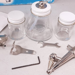 Peut inclure: Un ensemble d'outils d'a&eacute;rographe comprenant un a&eacute;rographe argent&eacute;, un a&eacute;rographe rouge et argent&eacute;, une cl&eacute;, un support argent&eacute;, un petit outil argent&eacute; et trois bocaux en verre avec des couvercles blancs. Les bocaux sont &eacute;tiquet&eacute;s "Pasche".