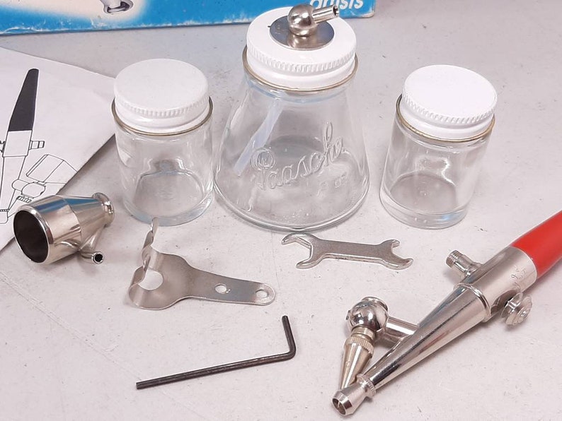 Peut inclure: Un ensemble d'outils d'a&eacute;rographe comprenant un a&eacute;rographe argent&eacute;, un a&eacute;rographe rouge et argent&eacute;, une cl&eacute;, un support argent&eacute;, un petit outil argent&eacute; et trois bocaux en verre avec des couvercles blancs. Les bocaux sont &eacute;tiquet&eacute;s "Pasche".