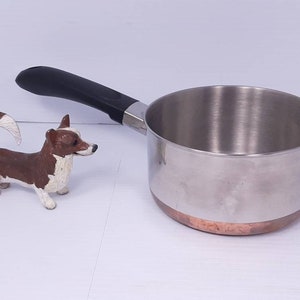 May include: A stainless steel saucepan with a copper bottom and a black handle. The saucepan is empty and sitting on a white surface.