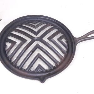May include: A dark brown, cast iron grill pan with a circular design and a handle. The pan features a geometric pattern of angled lines within the cooking surface. The handle is curved for easy grip.