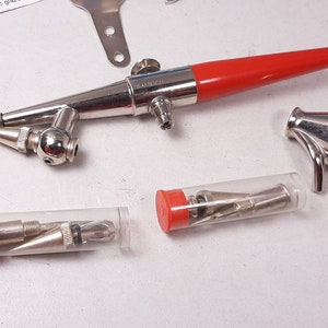 Peut inclure: Un a&eacute;rographe argent&eacute; avec une poign&eacute;e rouge et une buse argent&eacute;e. L'a&eacute;rographe est accompagn&eacute; d'une petite tasse argent&eacute;e et de trois tubes contenant des aiguilles de diff&eacute;rentes tailles.