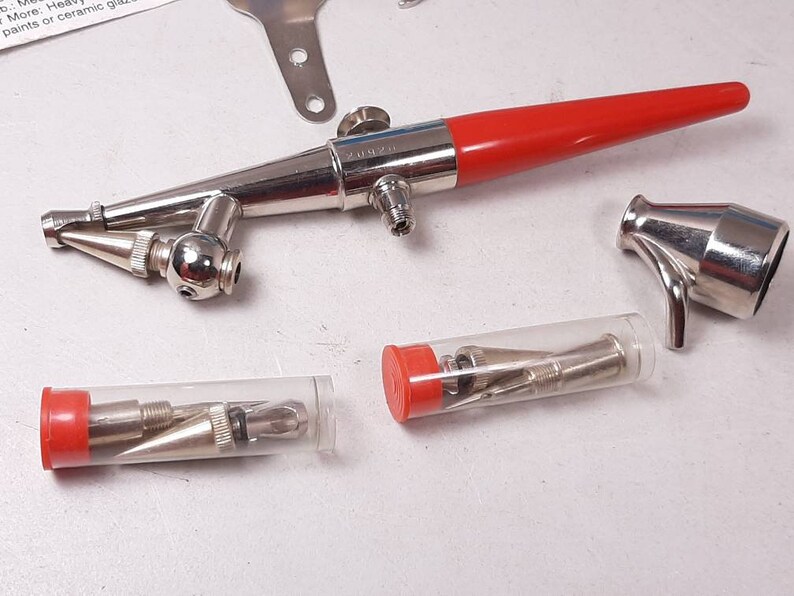 Peut inclure: Un a&eacute;rographe argent&eacute; avec une poign&eacute;e rouge et une buse argent&eacute;e. L'a&eacute;rographe est accompagn&eacute; d'une petite tasse argent&eacute;e et de trois tubes contenant des aiguilles de diff&eacute;rentes tailles.