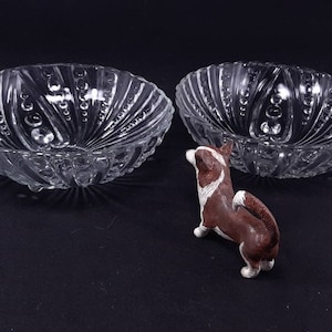 May include: Two clear glass bowls with a textured, swirled design and bubble accents. A small brown and white dog figurine is positioned between the bowls. The bowls are likely for serving or display.