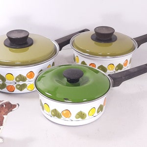 May include: A set of three vintage enamel saucepans with lids. The pans have white bodies with a colorful leaf and fruit pattern. The lids are olive green and dark green, and the handles are black.
