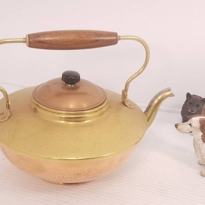 May include: A gold and copper kettle with a wooden handle. The kettle has a round body and a spout on the side. The kettle is sitting on a white surface.