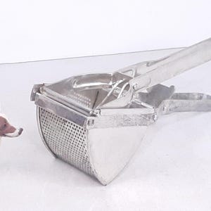 May include: A silver potato ricer with a handle and a perforated base. The ricer is made of metal and has a simple design.