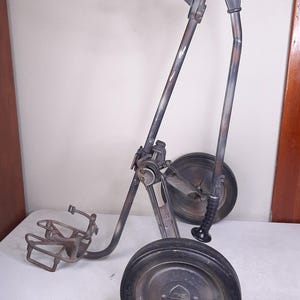 May include: A vintage golf bag push cart with two large black wheels and a metal frame. The cart has a handle and a bracket for holding a golf bag. The metal frame has a faded, mottled paint finish.