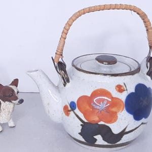 May include: A white ceramic teapot with a brown bamboo handle and floral design. The teapot has a lid and a spout. The flowers are orange, blue, and black.
