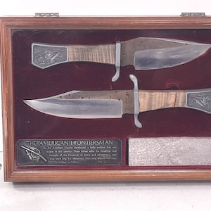 May include: Two antique-style knives with wooden handles are displayed in a wooden shadow box with a velvet lining. The knives are displayed on a red velvet background. The shadow box has a plaque with the text "The American Frontiersman" and a description of the American frontiersman.