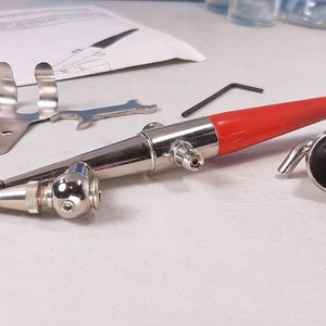 Peut inclure: Un a&eacute;rographe argent&eacute; avec une poign&eacute;e rouge et une buse argent&eacute;e. L'a&eacute;rographe est sur une surface blanche avec une cl&eacute; &agrave; molette argent&eacute;e et un support argent&eacute;.