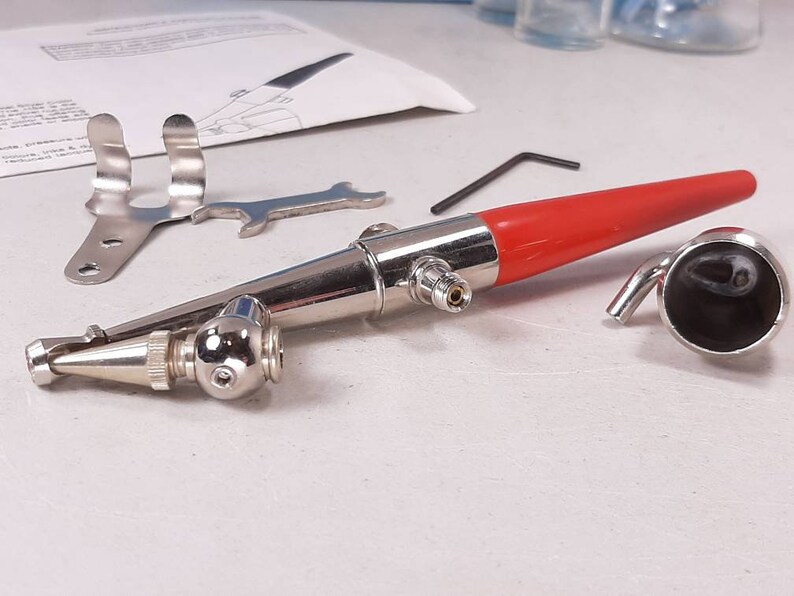 Peut inclure: Un a&eacute;rographe argent&eacute; avec une poign&eacute;e rouge et une buse argent&eacute;e. L'a&eacute;rographe est sur une surface blanche avec une cl&eacute; &agrave; molette argent&eacute;e et un support argent&eacute;.