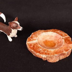 May include: A brown and white ceramic dog figurine next to a round, orange-brown, natural stone ashtray.