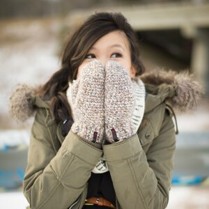 May include: A woman wearing a green parka with a fur-trimmed hood, covering her face with a pair of brown and white knitted gloves.