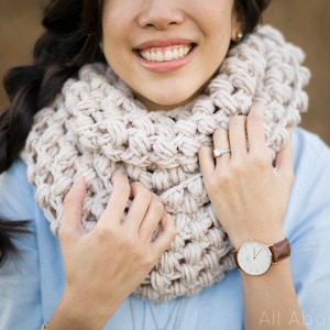 May include: A woman wearing a light blue shirt with a white crocheted infinity scarf. She is holding the scarf in front of her and smiling. She is wearing a watch on her left wrist.