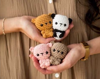 May include: Four small crocheted stuffed animals in the shape of bears and a panda. The animals are in a person's hands. The bears are brown, pink, and tan. The panda is white and black.
