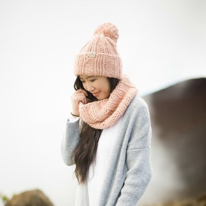 May include: A woman wearing a pink knitted hat with a pom pom and a matching scarf. She is wearing a gray cardigan and a white shirt. She is standing in front of a rocky background.