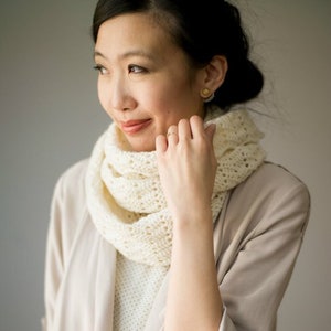 May include: A woman wearing a beige jacket and a white crocheted infinity scarf. The scarf is loosely draped around her neck and she is holding it with her right hand.