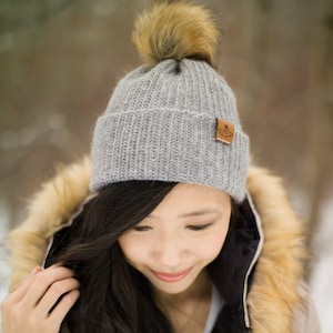 Pode incluir: Uma mulher usando um gorro de tricô cinza com um pompom de pele sintética marrom, um casaco de inverno rosa com uma borda de pele sintética e cabelo comprido e escuro.