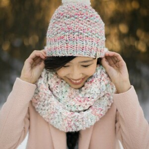 May include: A woman wearing a pink coat, a white pom-pom beanie with a multi-colored knit pattern, and a matching multi-colored infinity scarf.