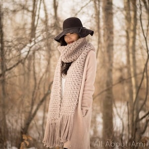 May include: A woman wearing a wide-brimmed black hat and a long, light pink crocheted scarf with fringe. She is standing in a snowy forest.