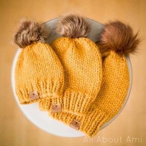 May include: Three yellow knitted hats with brown faux fur pom-poms. Each hat has a small brown leather tag with a floral design.