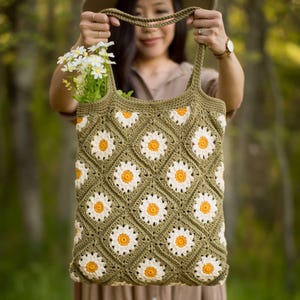 May include: Olive green crochet tote bag with a daisy pattern. The bag features white and yellow daisy motifs arranged in a diamond pattern. The bag is held up by a person. The bag is filled with white flowers.