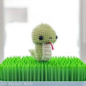 Puede incluir: Una serpiente de crochet verde con lengua roja y ojos negros, sentada sobre una superficie verde como hierba.